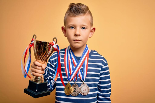 Young little caucasian kid wearing winner medals and victory award trophy over yellow background with a confident expression on smart face thinking serious - Foto, immagini