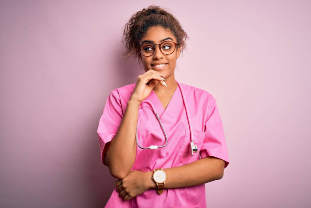 Африканська дівчинка-медсестра в медичній формі і стетоскоп над рожевим фоном з рукою на підборіддя думати про питання, задумливий вираз. Посміхаючись із задумливим обличчям. Концепція сумнівів. - Фото, зображення