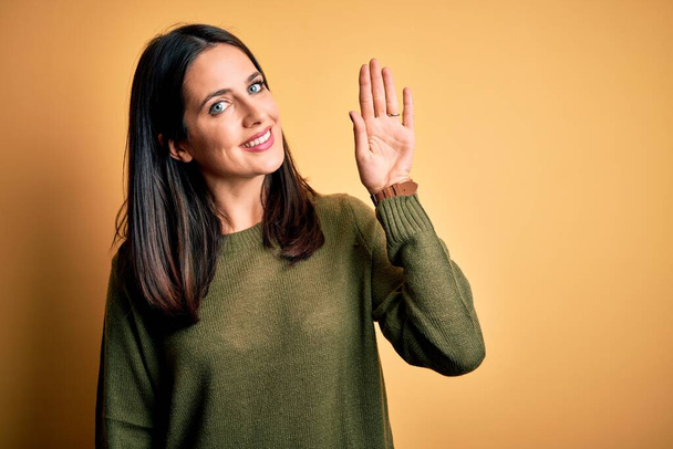 Young brunette woman with blue eyes wearing green casual sweater over yellow background Waiving saying hello happy and smiling, friendly welcome gesture - Valokuva, kuva