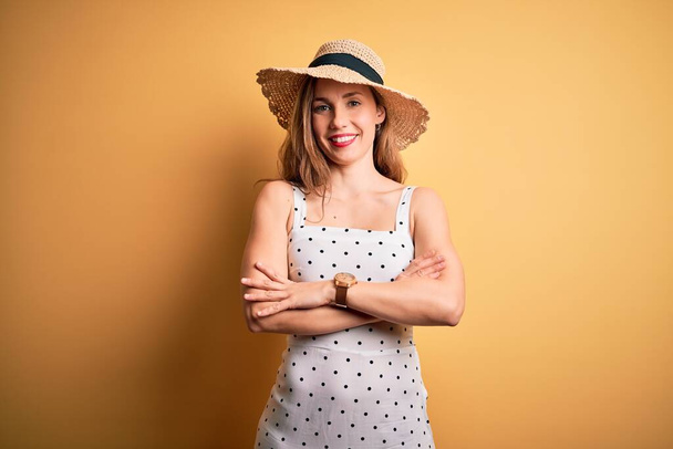 Mladá krásná blondýnka na dovolené na sobě letní klobouk přes žluté pozadí šťastný obličej s úsměvem se zkříženými pažemi dívá do kamery. Pozitivní osoba. - Fotografie, Obrázek