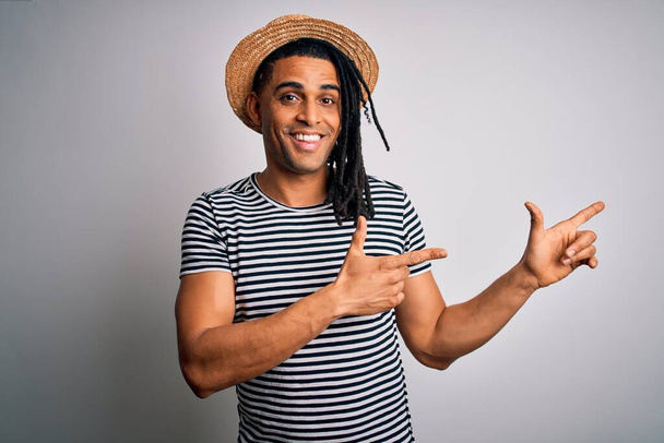 Jovem afro-americano com dreadlocks em férias vestindo camiseta listrada e chapéu sorrindo e olhando para a câmera apontando com duas mãos e dedos para o lado
. - Foto, Imagem