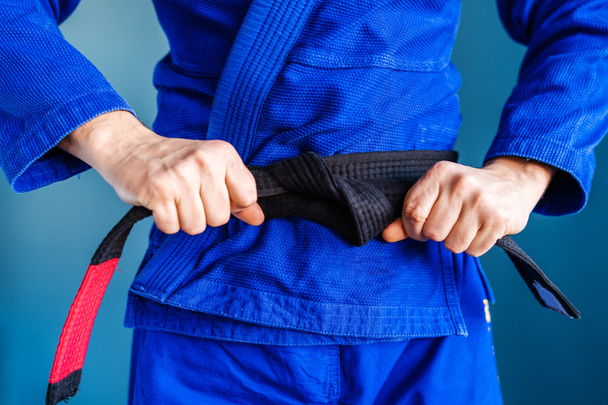 Nahaufnahme auf dem Mittelteil des bjj Brazilian Jiu Jitsu Black Gürtel Hände halten und Krawatte um die Taille eines Athleten Kämpfer tragen blauen Kimono Gi steht vor der Wand - Foto, Bild