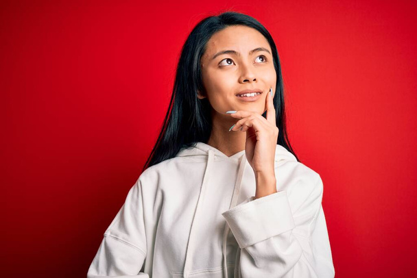 Young beautiful chinese sporty woman wearing sweatshirt over isolated red background with hand on chin thinking about question, pensive expression. Smiling with thoughtful face. Doubt concept. - Photo, Image