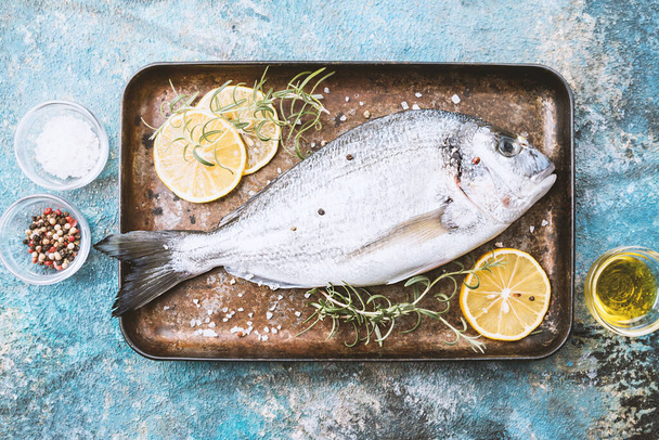 Fresh uncooked dorado or sea bream fish on tray with lemon slices and herbs over blue background, top view - Foto, Imagem