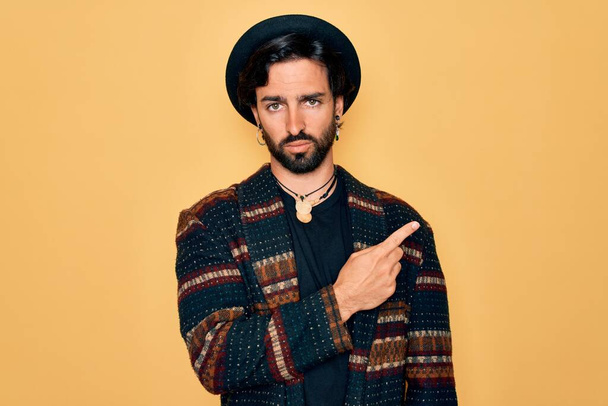 Jeune homme bohème hispanique beau portant le style hippie et chapeau boho Pointant avec le doigt de la main sur le côté montrant la publicité, visage sérieux et calme
 - Photo, image