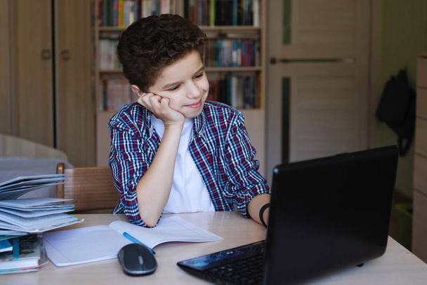 Tinédzser fiú egy asztalnál laptoppal belenéz a monitorba, és a kezével támasztja a fejét. A koronavírus karantén alá helyezte az otthoni oktatást. Szelektív fókusz. Homályos háttér. - Fotó, kép
