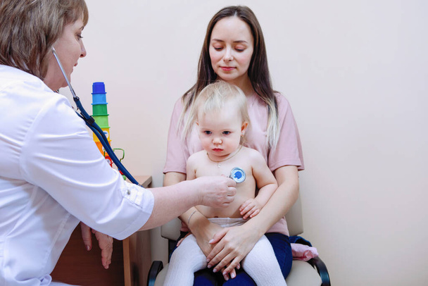 Gyermekgyógyászati tüdő légzésvizsgálat. Egy szőke kislány ül anya ölében. Női orvos egy orvosi klinikán. - Fotó, kép