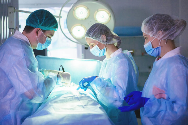 Három sebész a hasi műtét alatt. Kék egyenruha, gumikesztyű, orvosi műszerek. Helyszíni világítás. - Fotó, kép