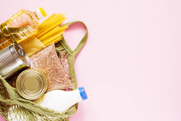 Food donations - oil, buckwheat, pasta, milk, canned food in a grocery grid on a pink background, top view with copy space. Food delivery concept. - 写真・画像