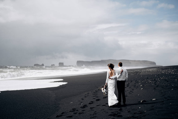Miejsce docelowe Islandia ślub. Para weselna spaceruje po czarnej plaży Vic. Piaszczysta plaża z czarnym piaskiem na wybrzeżu Oceanu Atlantyckiego. Ogromne piankowe fale. Pan młody przytula pannę młodą. - Zdjęcie, obraz