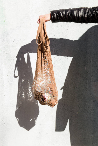 The delivery of food. Hand holding a mesh shopping bag with fresh food, hard shadows on the wall. Eco-friendly responsible lifestyle and shopping. A healthy diet, the concept of zero waste. - Fotografie, Obrázek