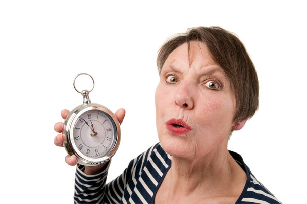Hoofd-en-schouder zicht van een oudere vrouw met een klok in haar rechterhand en bang met ronde mond kijkend naar de camera tegen een witte achtergrond. - Foto, afbeelding