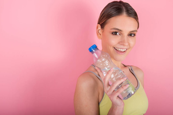 Kaunis istuvuus tyttö tilalla pullo vettä, hymyilee ja juo makeaa vettä, eristetty vaaleanpunainen tausta - Valokuva, kuva