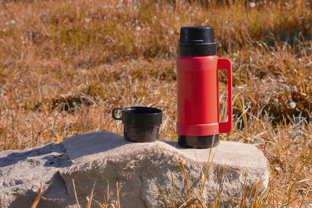 garrafa térmica vermelha com uma xícara em uma pedra na floresta no gramado
. - Foto, Imagem