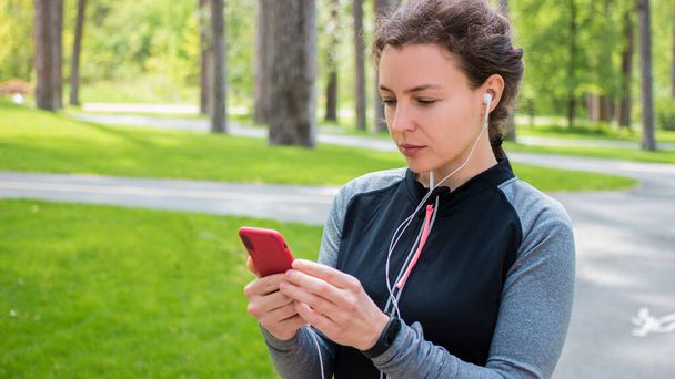 Young concentrated sporty woman in headphones holding smartphone and looking at screen,sharing running data on social media,listening music on mobile phone after jogging,training,work out  - Zdjęcie, obraz