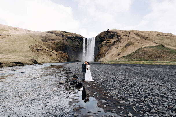 目的地アイスランドの結婚式。Skogafoss滝の近くの結婚式のカップル.花嫁と新郎が川の近くで抱き合っている. - 写真・画像
