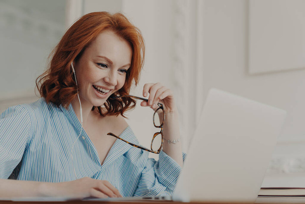 Positive redhead businesswoman participates in online conference, has training class with tutor, focused in display of laptop computer, holds spectacles, enjoys watching educational webinar. - Photo, Image