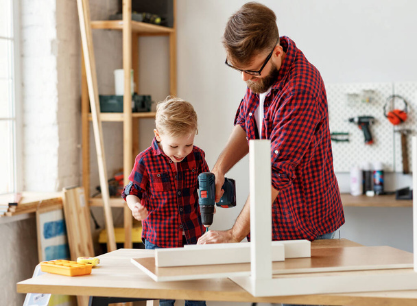 Opgewonden kleine jongen en jonge vader in soortgelijke shirts samen te houden boor terwijl het werken met hout in moderne handwerk studi - Foto, afbeelding