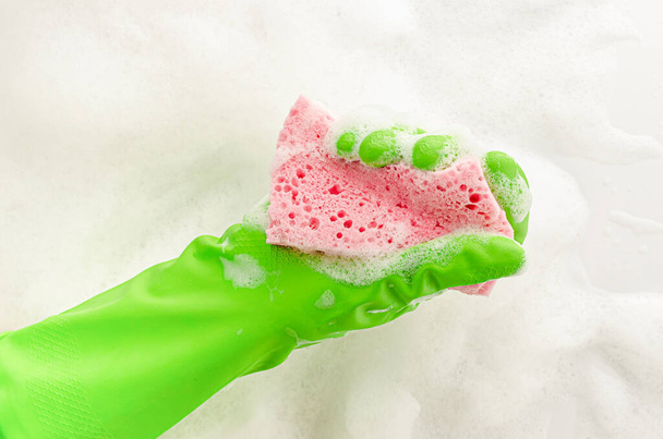 Ręka w rękawicy ochronnej trzymając gąbkę mydlaną na tle pianki. Mycie naczyń. Przestrzeń kopiowania - Zdjęcie, obraz