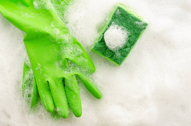 Vista superior de guantes verdes protectores y esponja sobre fondo jabonoso. Concepto de trabajo doméstico. Copiar espacio, vista superior
 - Foto, imagen