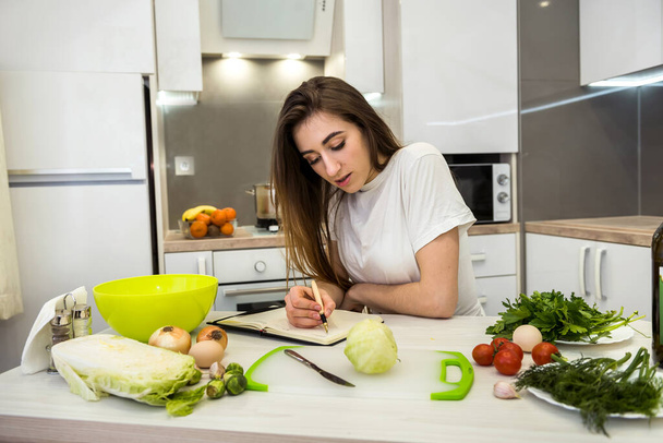 Junge hübsche Frau sitzt am Küchentisch mit verschiedenen Gemüsesorten und schreibt oder schreibt alles über gesunde Ernährung in ihr Tagebuch - Foto, Bild