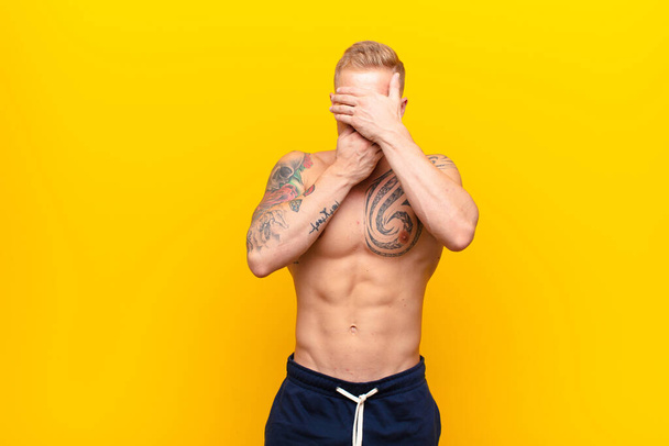young strong blonde man covering face with both hands saying no to the camera! refusing pictures or forbidding photos against yellow wall - Foto, Imagem