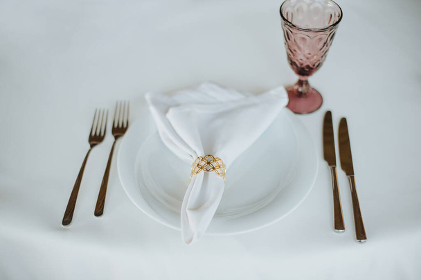 Il tavolo è coperto da una tovaglia bianca, servita. Piatti, posate, tovaglioli, bicchieri rosa, seggiolini, rametto di verdure
. - Foto, immagini