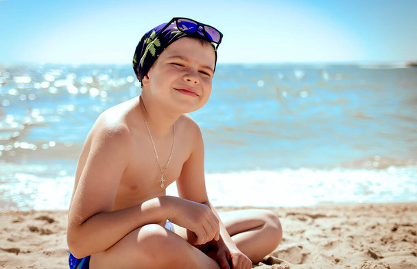 Un adolescent sourit et s'assoit sur le sable dans des maillots de bain. Le concept de détente et de tranquillité. Copiez l'espace sur la droite. Concept d'été et vacances
 - Photo, image
