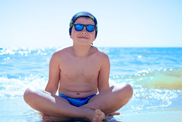 Teini-ikäinen poika istuu hiekalla uimahousuissa, sinisissä aurinkolaseissa, harjoittaa meditaatiota, rentoutumista ja joogaa meren rannalla. Rentoutumisen ja rauhallisuuden käsite. Kopioi tila - Valokuva, kuva