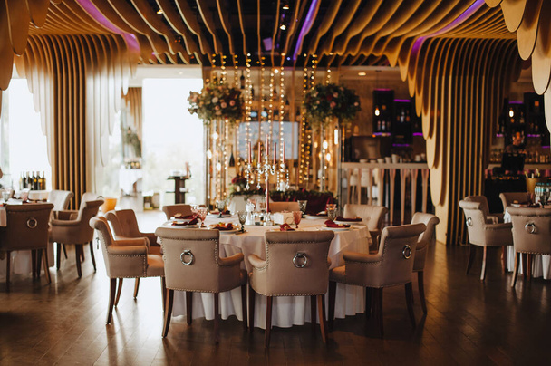 Modern wooden background, garland, interior. The tables are served, compositions of flowers and greenery. Plates, cutlery, napkin, pink glasses. Banquet hall. - Photo, Image