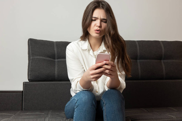 young beautiful brunette smiling womansitting on the sofa in her appartment looks amazed while replying to message in her smartphone, feeling cozy at home. - Φωτογραφία, εικόνα
