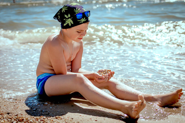Concept d'été et vacances. Adolescent garçon tient de nombreux coquillages de mer dans ses mains dans des maillots de bain et un bandana avec des lunettes bleues sur la mer. Copier l'espace sur la droite
 - Photo, image