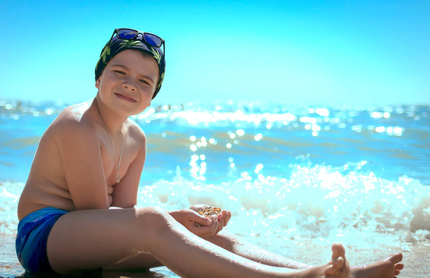 Teenager boy looking at the camera with a smile holds many sea shells in his hands in swimming trunks and a bandana with blue glasses on the sea. Summer and Vacations concept. Copy space on the right - Photo, Image