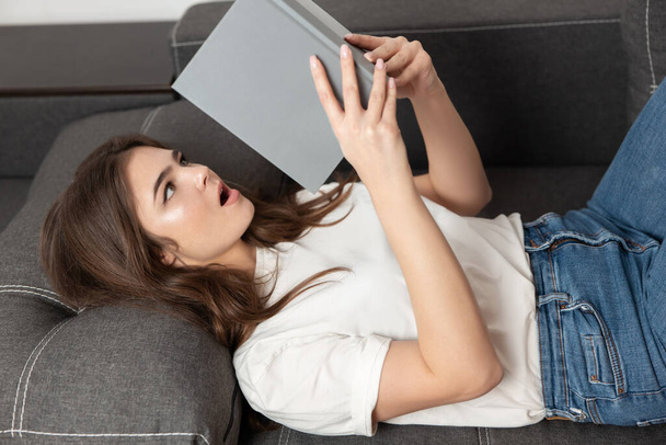 young beautiful brunette woman reading exciting book while lying on the sofa in her appartment looks very passionate, feeling cozy at home. - Fotografie, Obrázek