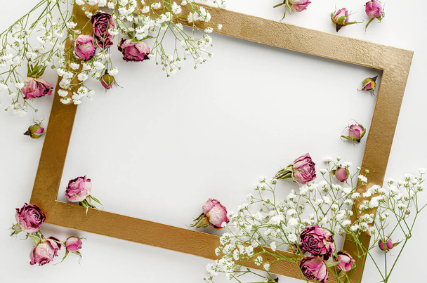 Creative golden frame decorated with dried pink roses on white background. Greeting card concept. Flat lay, copy space - Photo, Image