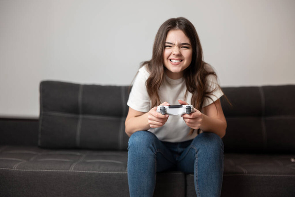 brunette young woman looks happy sitting on the sofa with joystick in her hands playing online game after hard work day ,feeling cozy at home. - Photo, Image