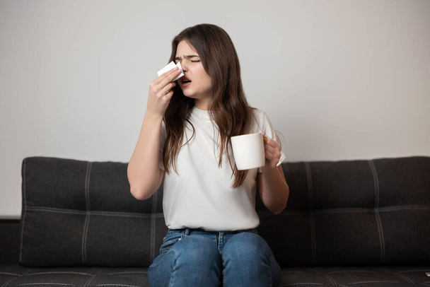 young beautiful brunette woman sits on the sofa bowing her running nose drinking medicine looking sick, healthcare and medical concept. - Foto, afbeelding