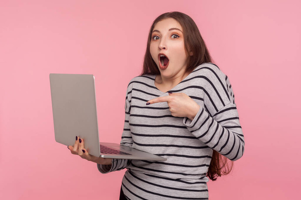 Wow, unbelievable! Portrait of shocked young woman in striped sweatshirt standing with open mouth and pointing at laptop, expressing great surprise. indoor studio shot isolated on pink background - Fotó, kép