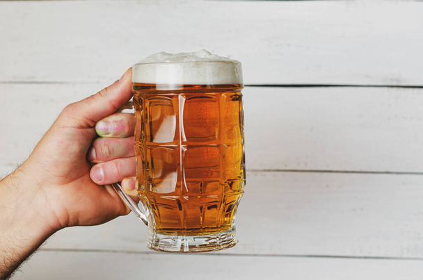 Male hand holding a full beer mug on white wooden background. Toned image. Copy space - Foto, Imagem