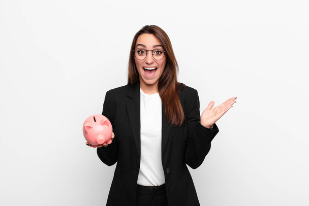 young businesswoman feeling happy, excited, surprised or shocked, smiling and astonished at something unbelievable with a piggy bank - Фото, изображение