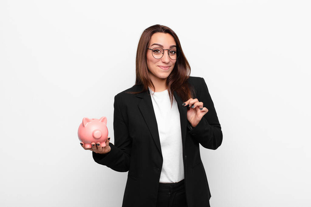 junge Geschäftsfrau, die arrogant, erfolgreich, positiv und stolz wirkt und mit einem Sparschwein auf sich selbst zeigt - Foto, Bild