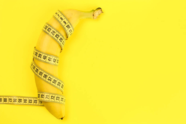 Banana wrapped with a tape measure on a yellow background, a concept of weight loss, healthy nutrition. Copy space - Fotografie, Obrázek