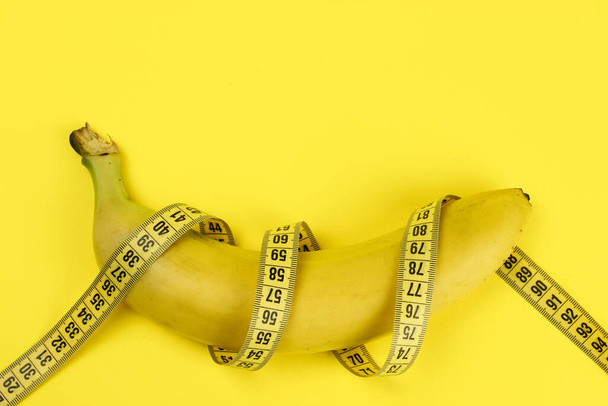 Banana wrapped with a tape measure on a yellow background, a concept of weight loss, healthy nutrition - Photo, Image