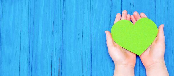 Top view of green heart in hands of child on wooden blue background. Copy space. Banner for site. - 写真・画像