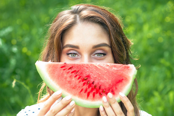 Young woman is covering her face by slice of watermelon on background of green grass. - Foto, imagen