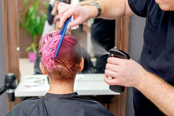 Vue arrière des mains du coiffeur ou du coiffeur vaporisant de l'eau sur les cheveux roses du client dans le studio de coiffure
. - Photo, image