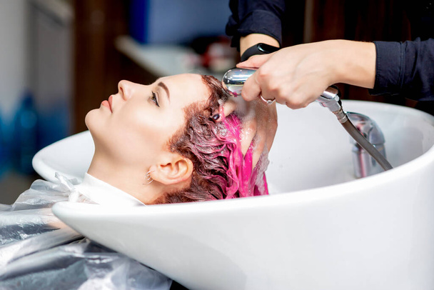 Widok z boku rąk fryzjera zmywanie farby z włosów kobiety w białym zlewie w salonie fryzjerskim. - Zdjęcie, obraz