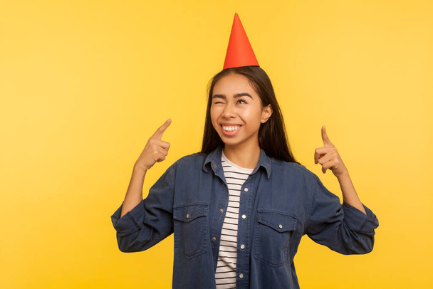Portrait of carefree excited girl in denim shirt pointing at funny party cone on head and smiling, celebrating successful event, birthday or holiday festival. studio shot isolated on yellow background - Photo, image