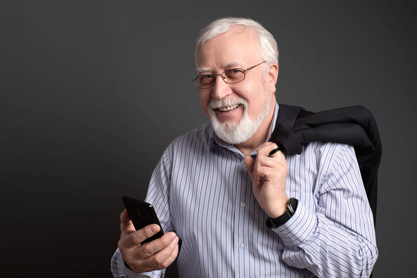 geschäftstüchtiger, weißhaariger Mann mit Brille und Telefon auf grauem Hintergrund lächelt freundlich - Foto, Bild