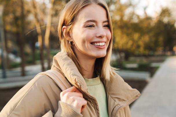 Ritratto di giovane donna bionda che indossa una giacca sorridente e guarda da parte mentre cammina nel parco
 - Foto, immagini